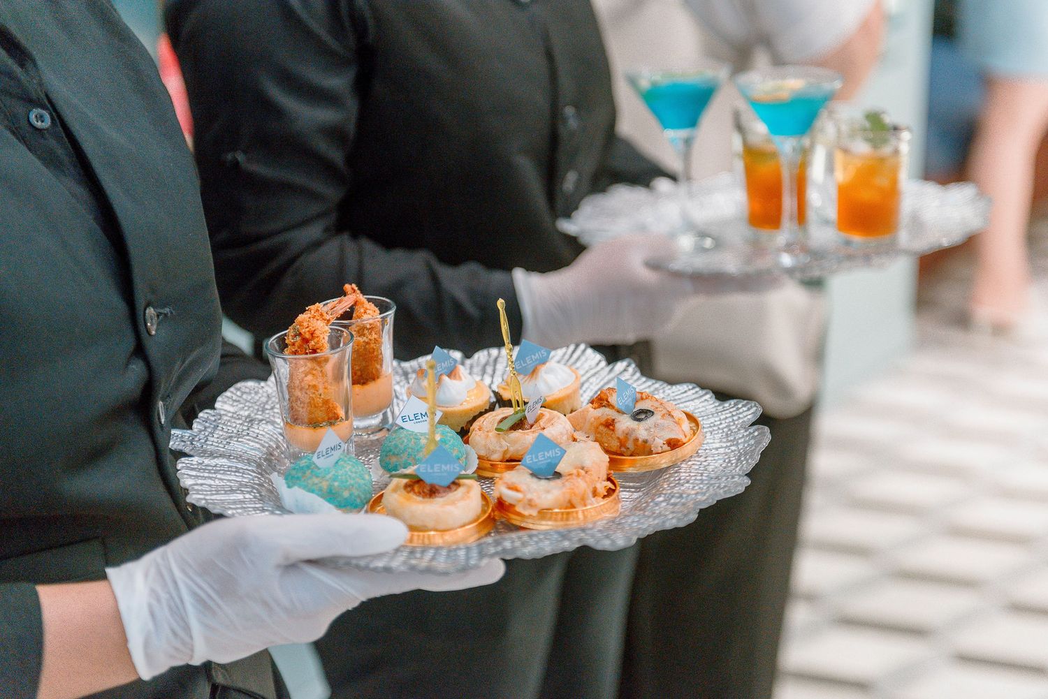 A server wearing white gloves carries a tray of appetizers with shrimp cocktails and blue cocktails in the background.