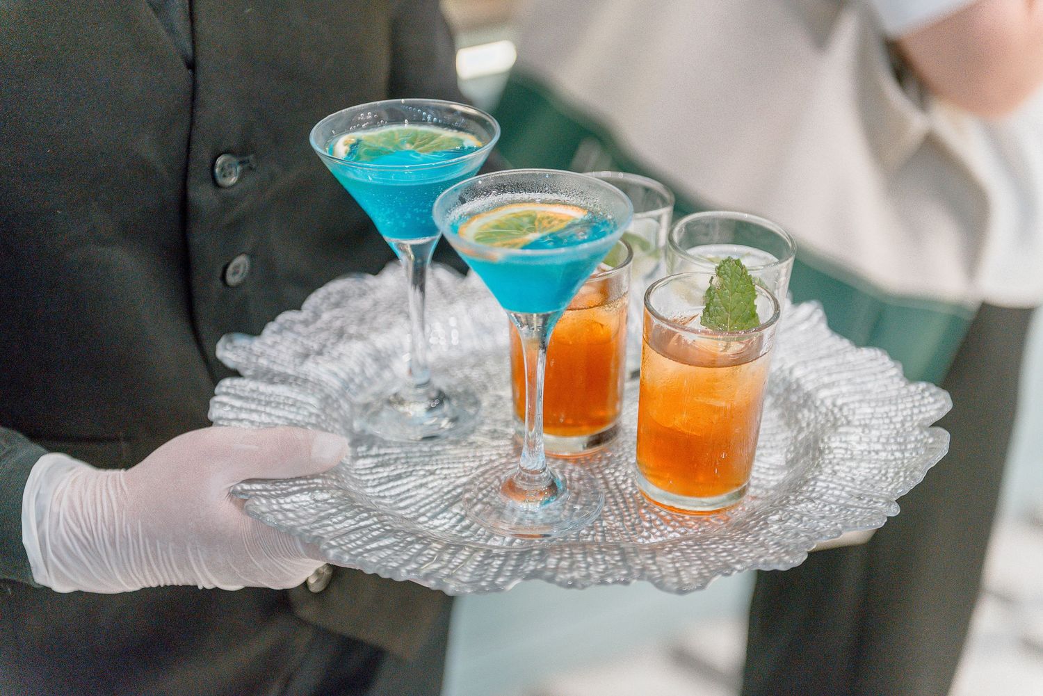 A server wearing white gloves holds a crystal tray with vibrant blue martinis and orange cocktails garnished with citrus.