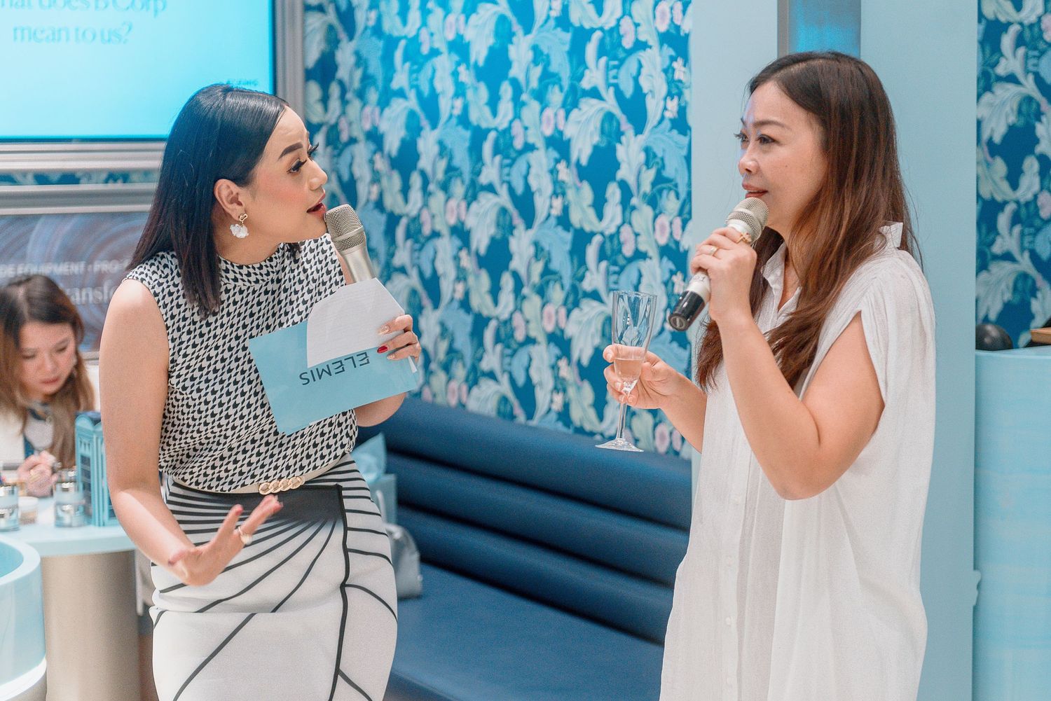 Two people having an animated conversation in front of a blue patterned wall in an interior setting.