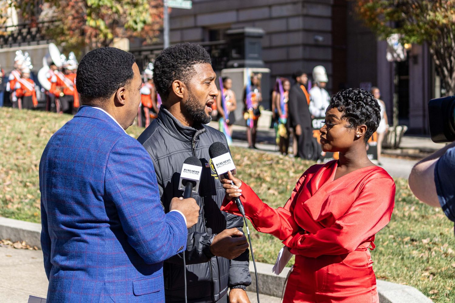 News reporters with microphones conduct an outdoor interview on a sunny day near a brick building.
