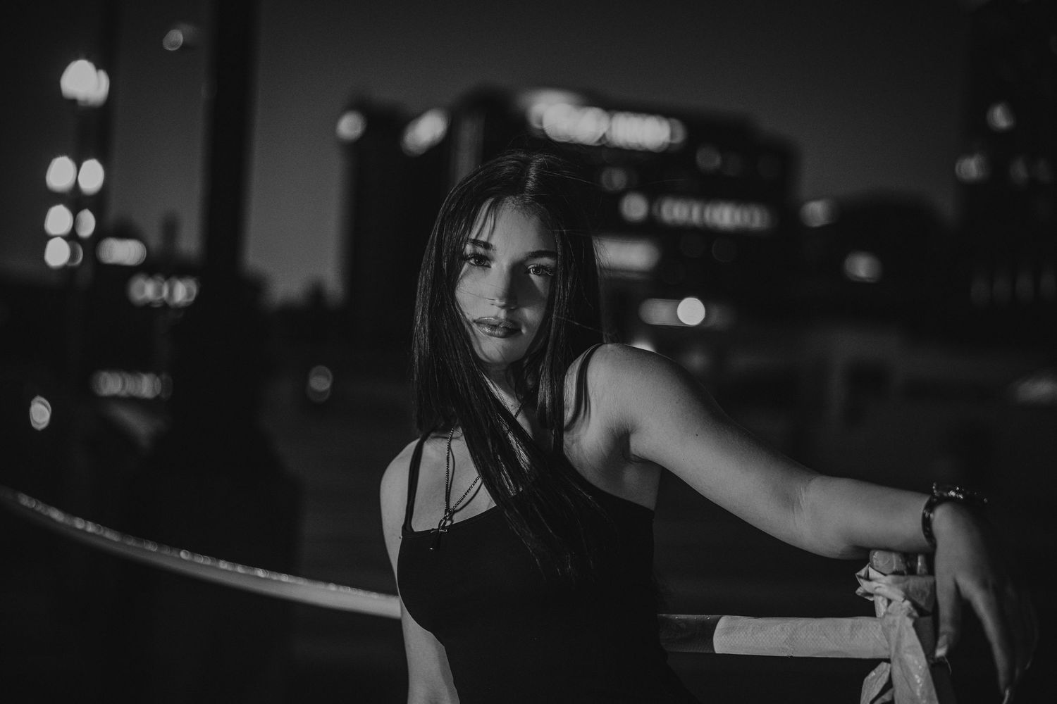 A black and white portrait of a woman in a tank top leaning against a railing at night with city lights in the background.