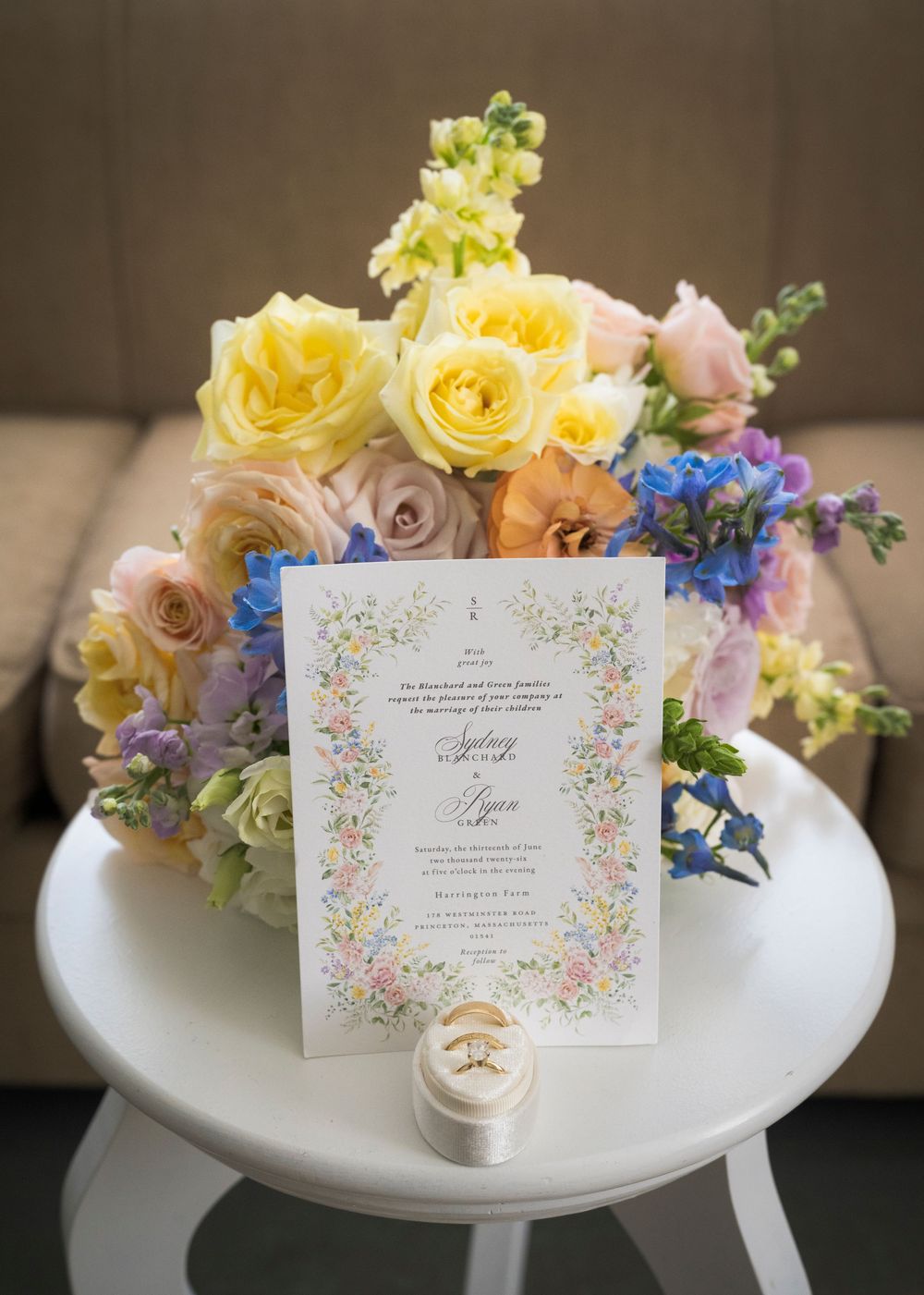 Colorful spring wedding invitation with floral border displayed beside pastel roses and blue flowers with gold rings.
