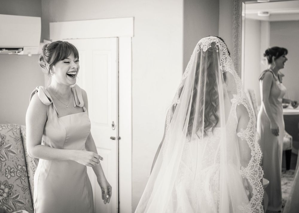 Black and white photo of a laughing bridesmaid helping a bride in a lace veil get ready before a wedding ceremony.