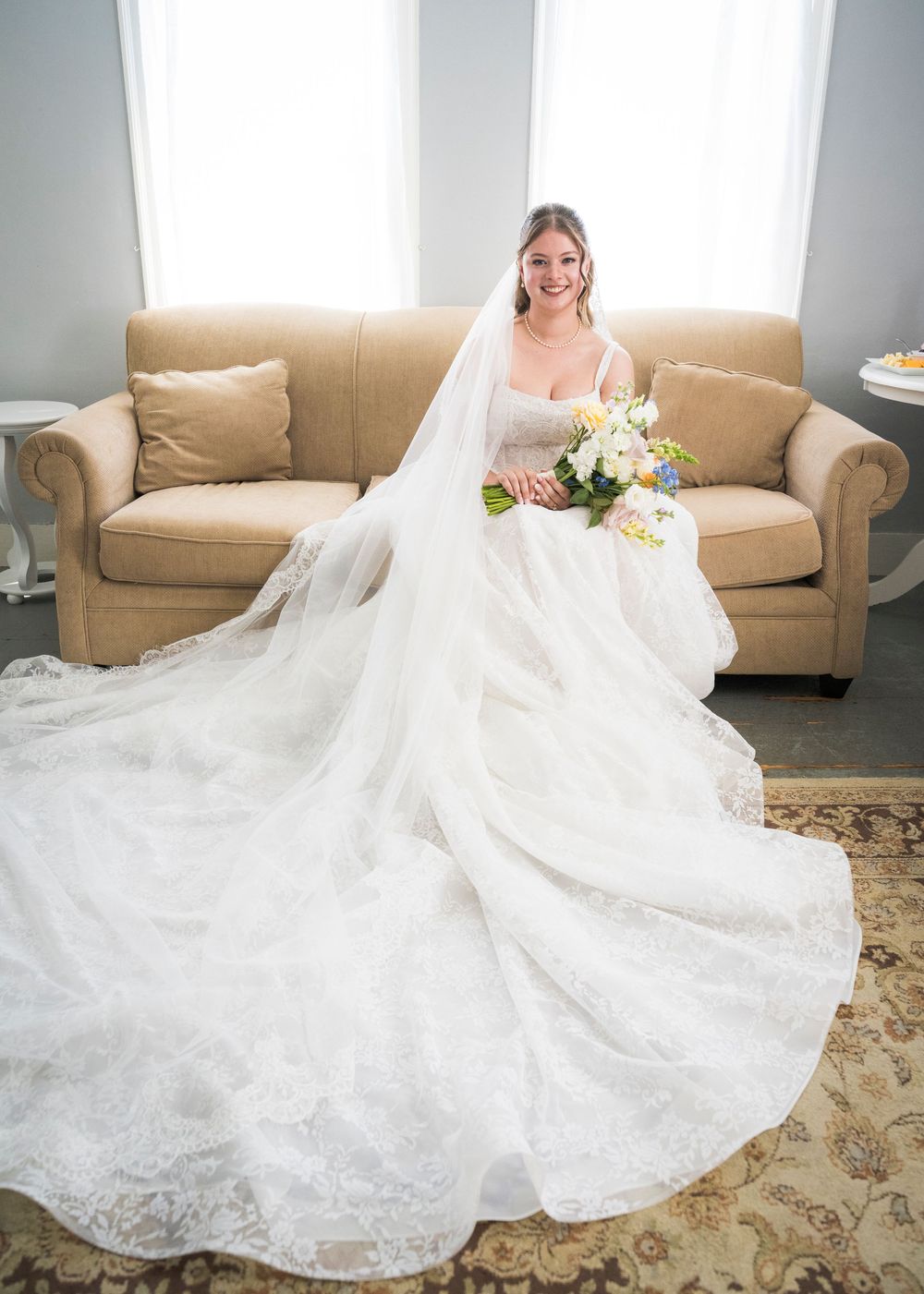 Smiling bride in elegant lace ball gown with cathedral veil seated on cream sofa holding white bouquet.