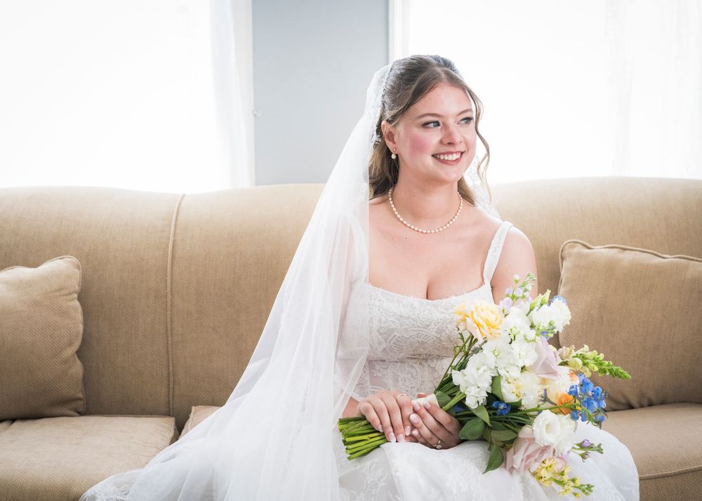 Beautiful bride in white gown holding colorful bouquet seated on beige sofa with veil and pearl jewelry.