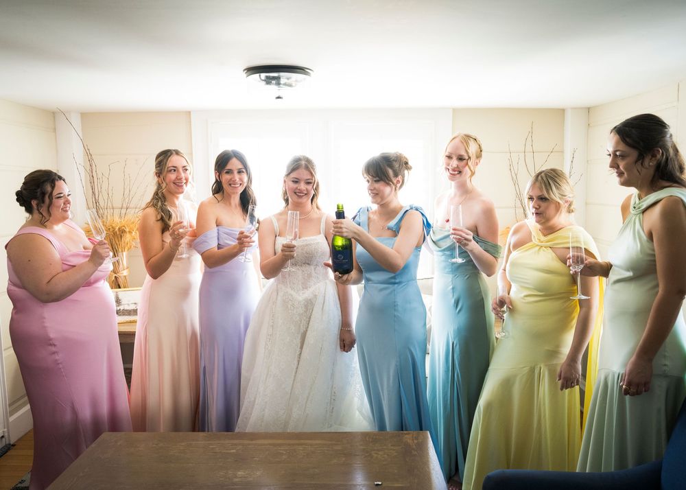 Bride and seven bridesmaids in rainbow pastel gowns celebrating with champagne bottle indoors.