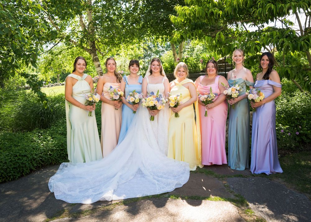 Bride poses with seven bridesmaids in pastel rainbow dresses holding bouquets in a lush garden.