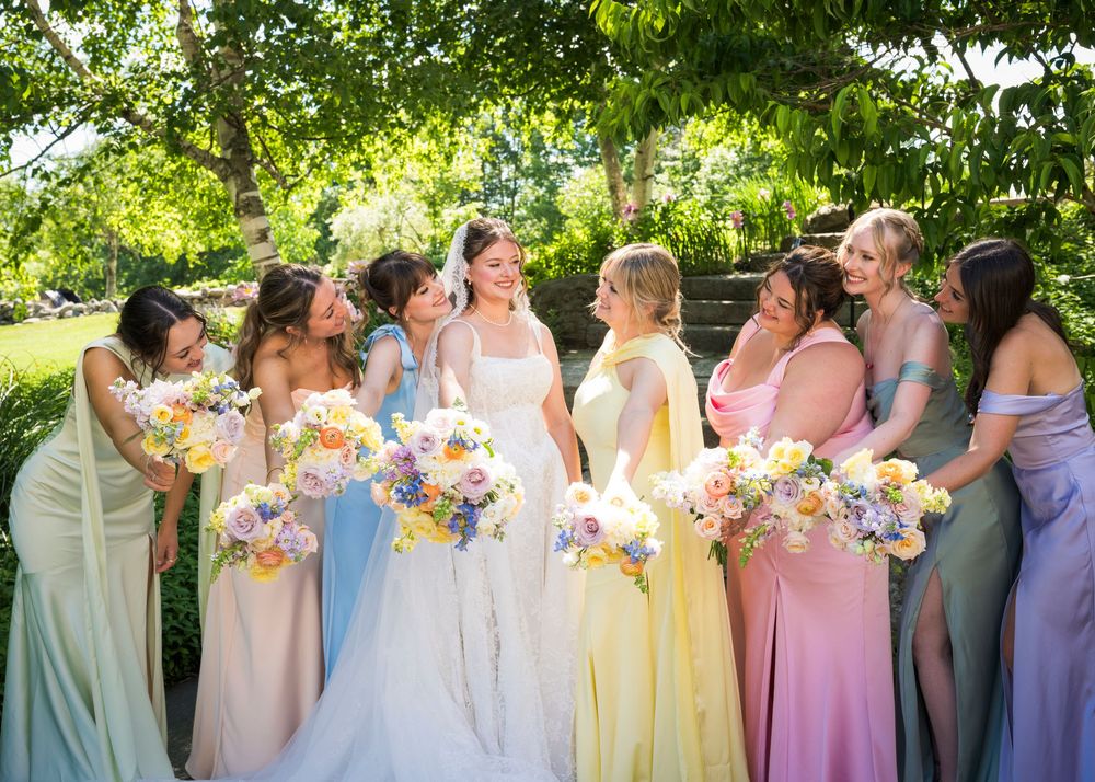 Bride surrounded by bridesmaids in colorful pastel gowns holding floral bouquets under garden trees.