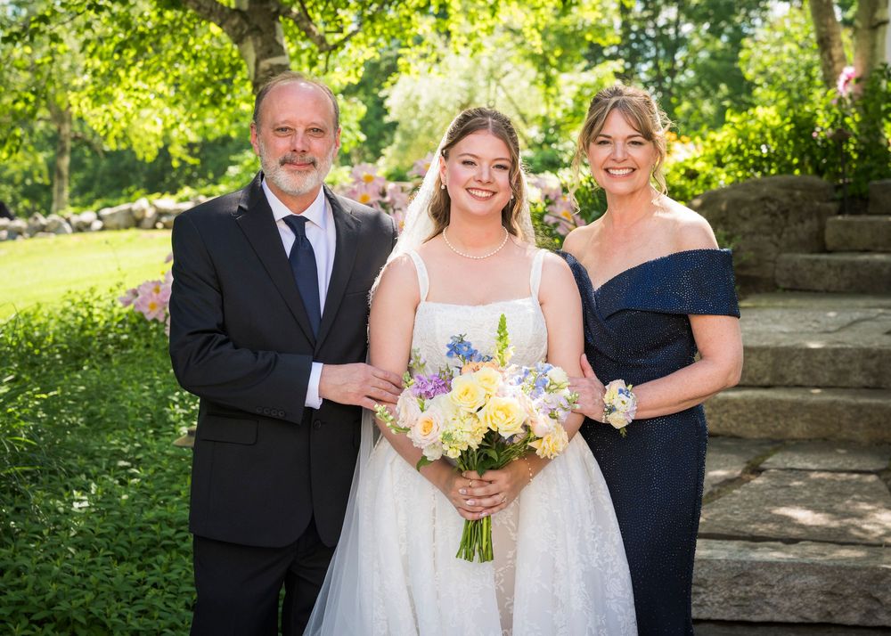 Bride holding colorful bouquet poses with parents in formal attire at outdoor garden wedding.
