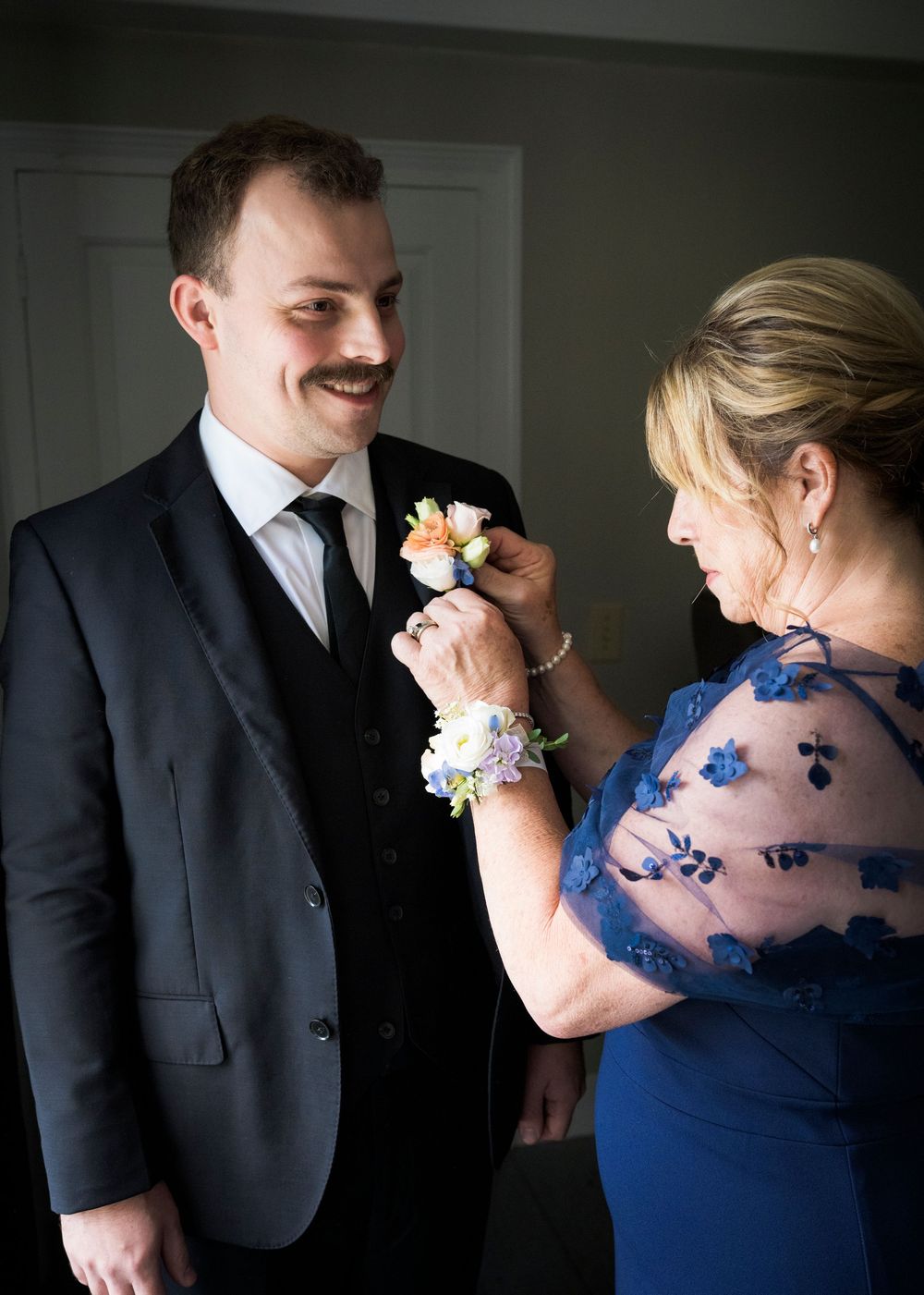 Groom smiles as woman in blue dress pins boutonniere on his dark suit lapel before wedding.