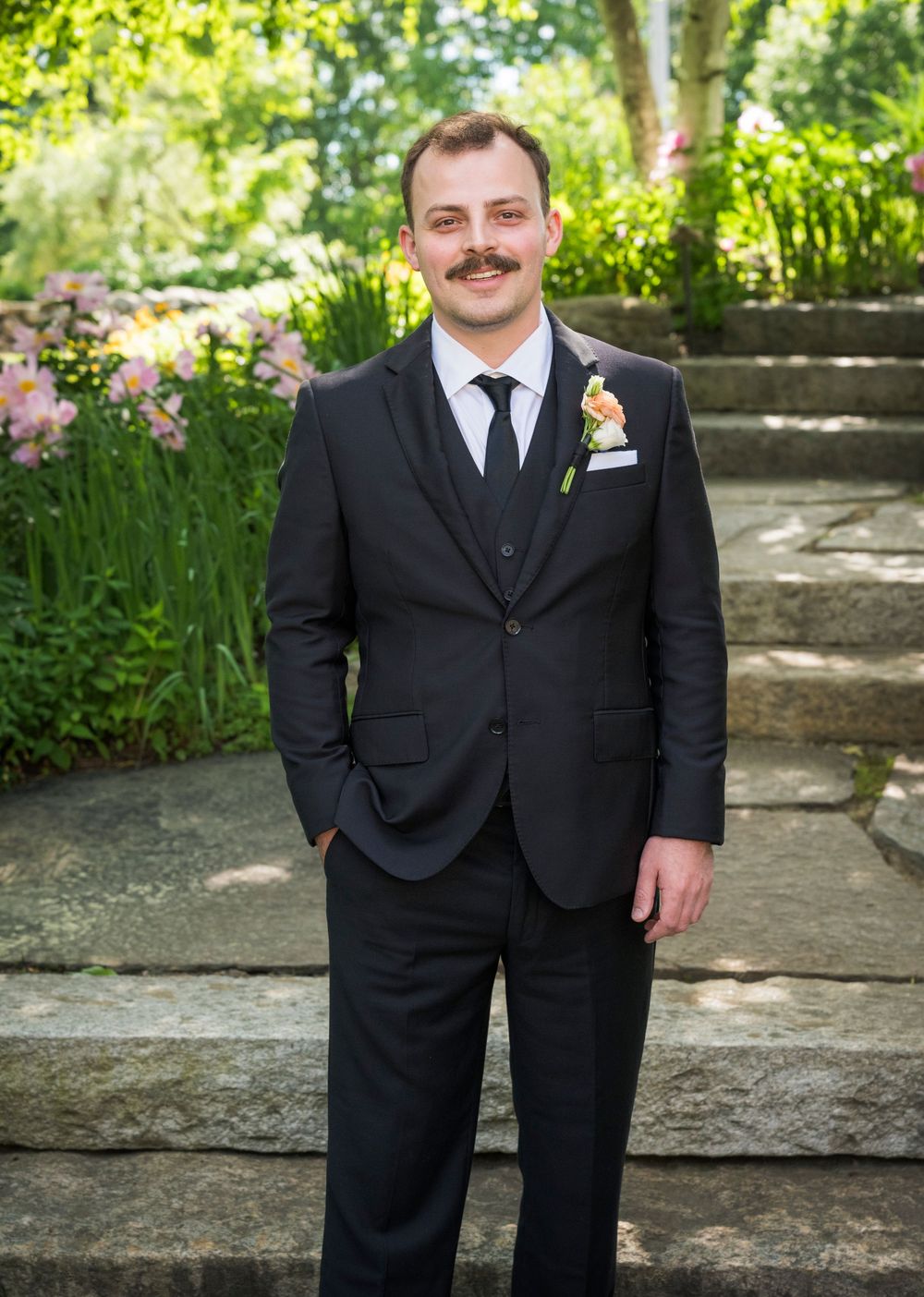 Dapper groom in a black three-piece suit with boutonniere standing on stone steps in a garden.
