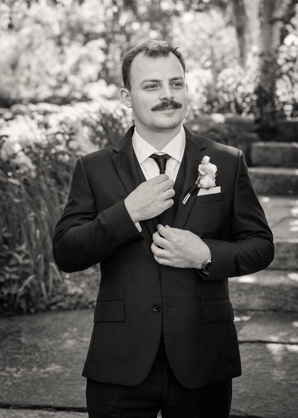 Dapper groom in black suit with boutonniere adjusting tie in black and white outdoor portrait.