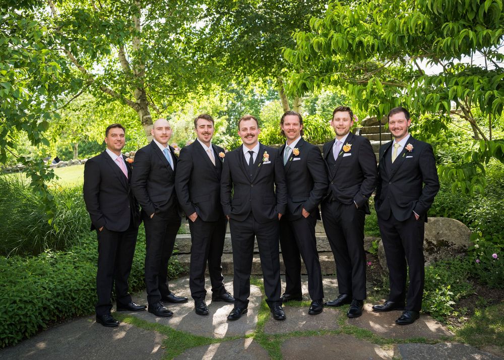 Groom and six groomsmen in matching dark suits with boutonnieres posing under green trees outdoors.