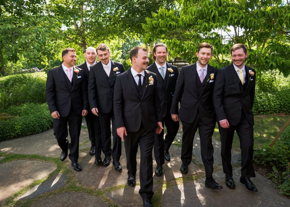 Groomsmen in black suits with boutonnieres walk together laughing in a lush garden setting.