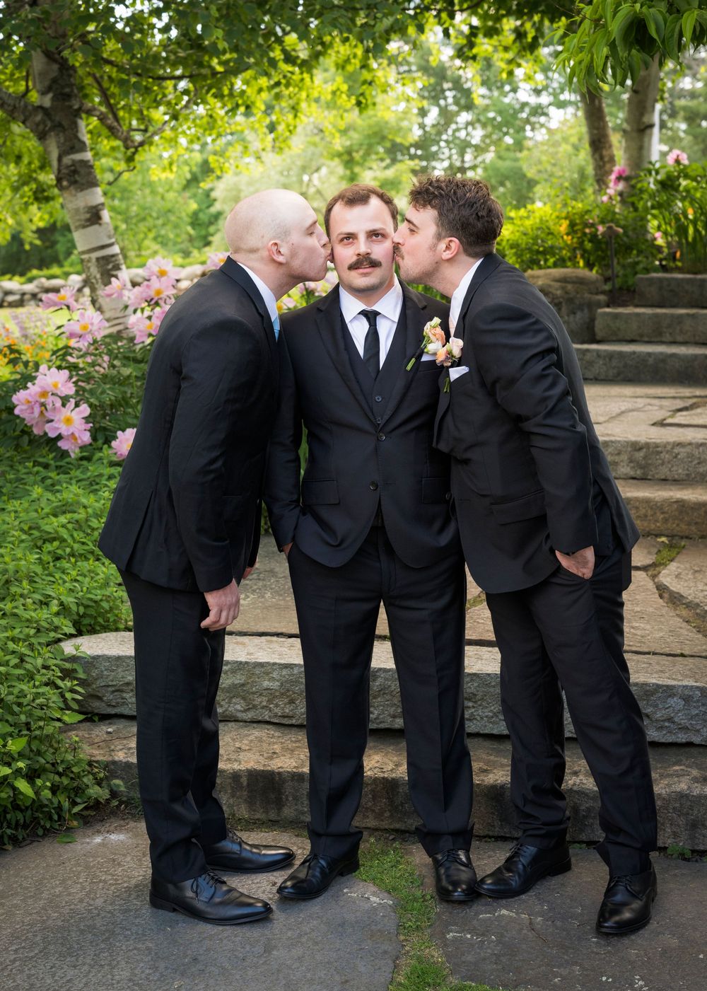 Groom with mustache receives kisses on cheeks from two groomsmen in black suits outdoors.