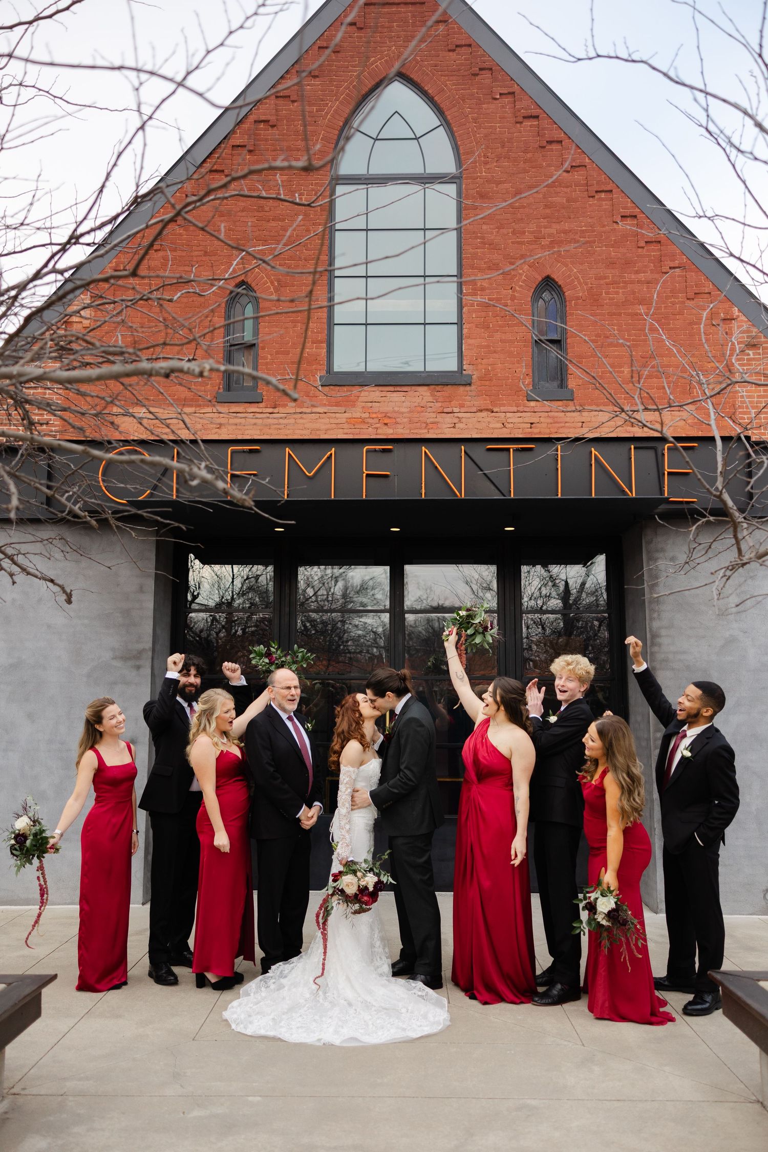 Wedding party celebrates outside Clementine venue with red bridesmaid dresses and formal attire in front of brick church facade.