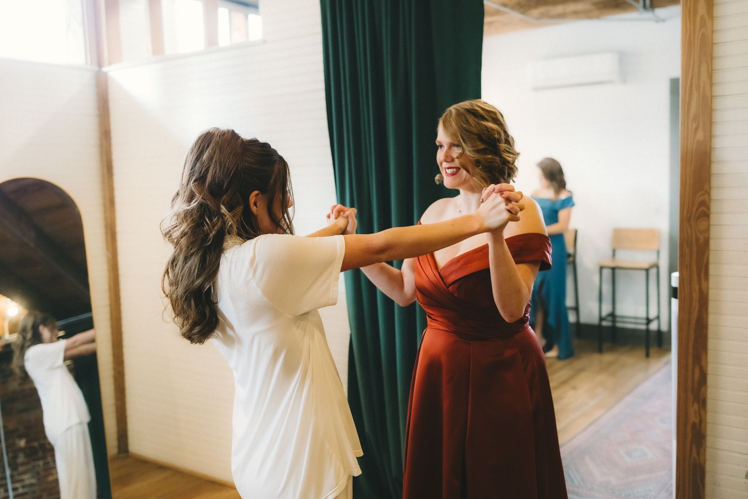 Dancers in white and burgundy outfits practice choreography in a bright studio with large windows.