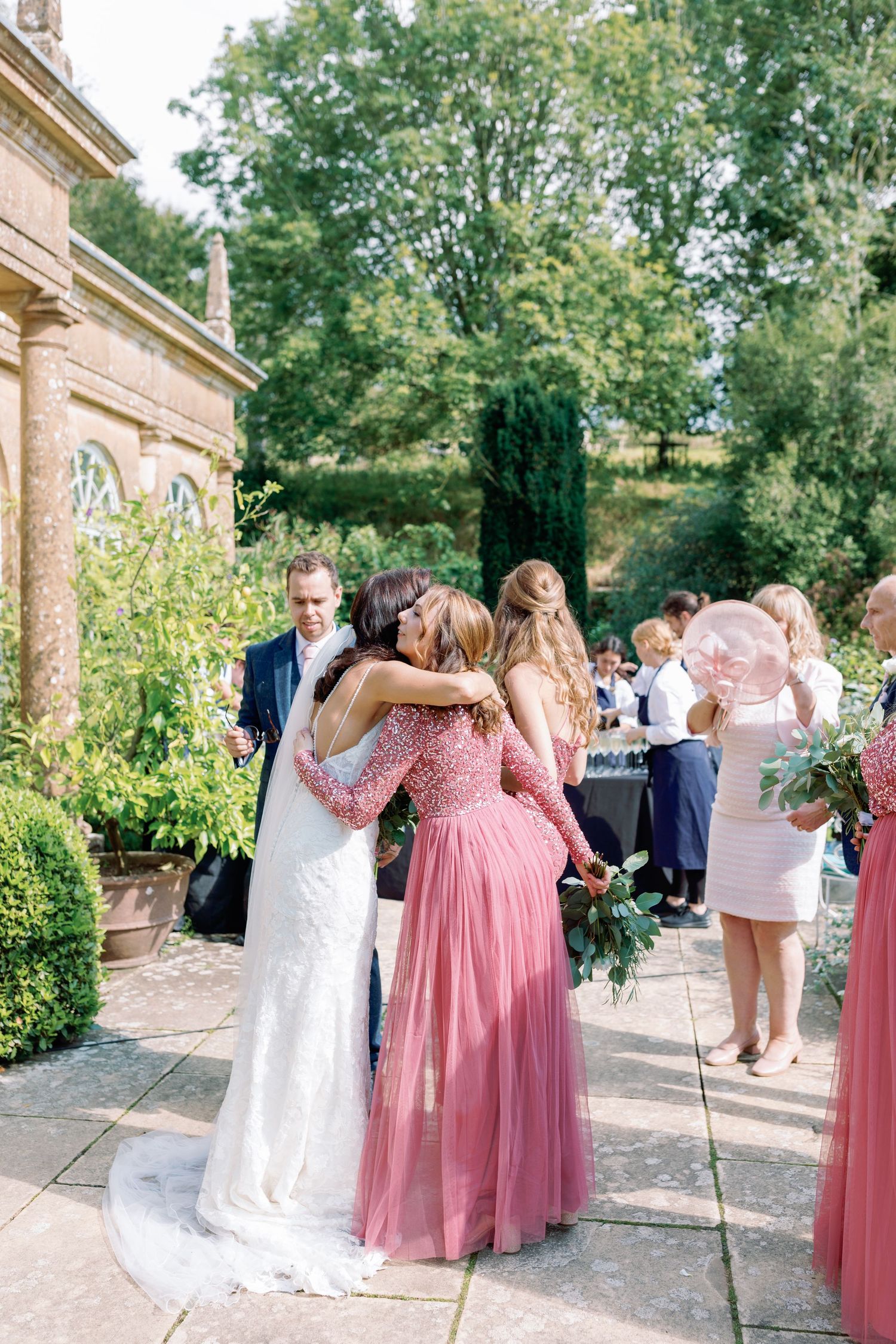 Wedding party members in pink bridesmaid dresses and white gowns embrace during an outdoor garden celebration.