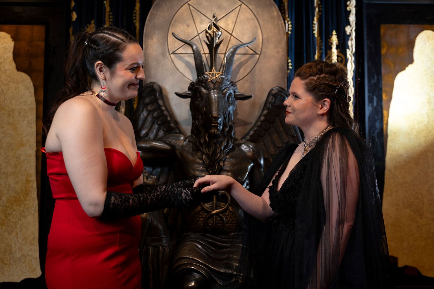 Two people in formal attire converse in front of an ornate gothic throne with a pentagram design in the background.