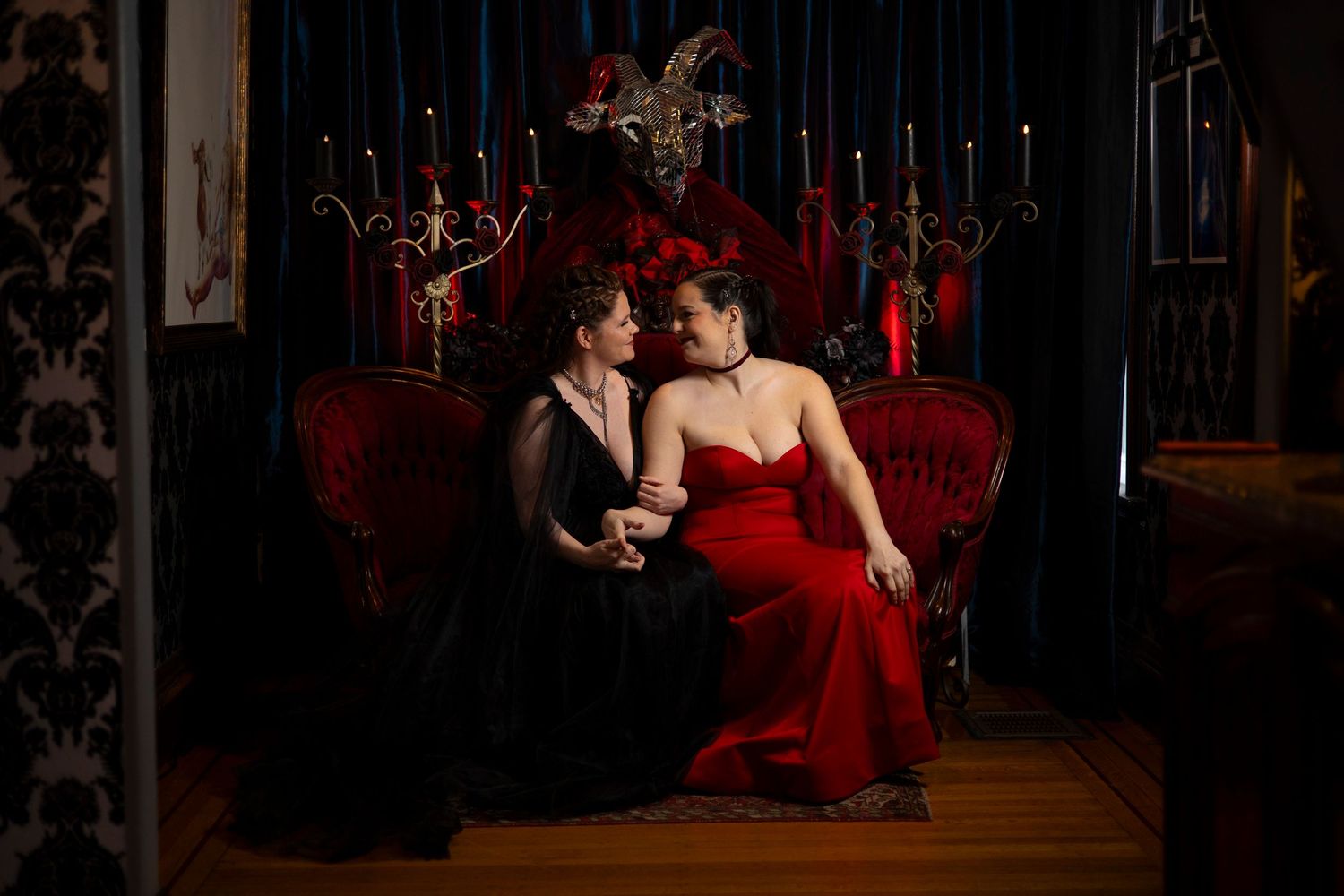 Elegant couple in formal attire poses intimately on a luxurious red velvet chaise lounge in a dimly lit studio setting.
