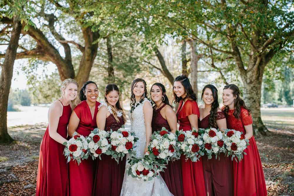 A wedding party in deep red bridesmaid dresses poses outdoors with white and red bouquets in a natural setting.