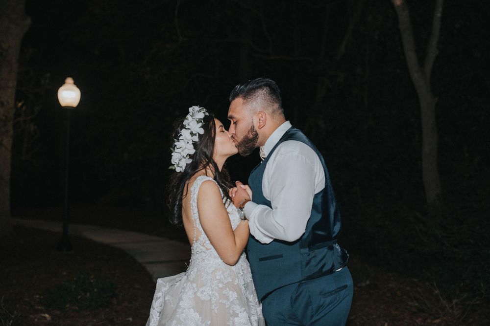 A couple shares a romantic kiss at night outdoors, with the bride in a lace dress and groom in a vest and tie.