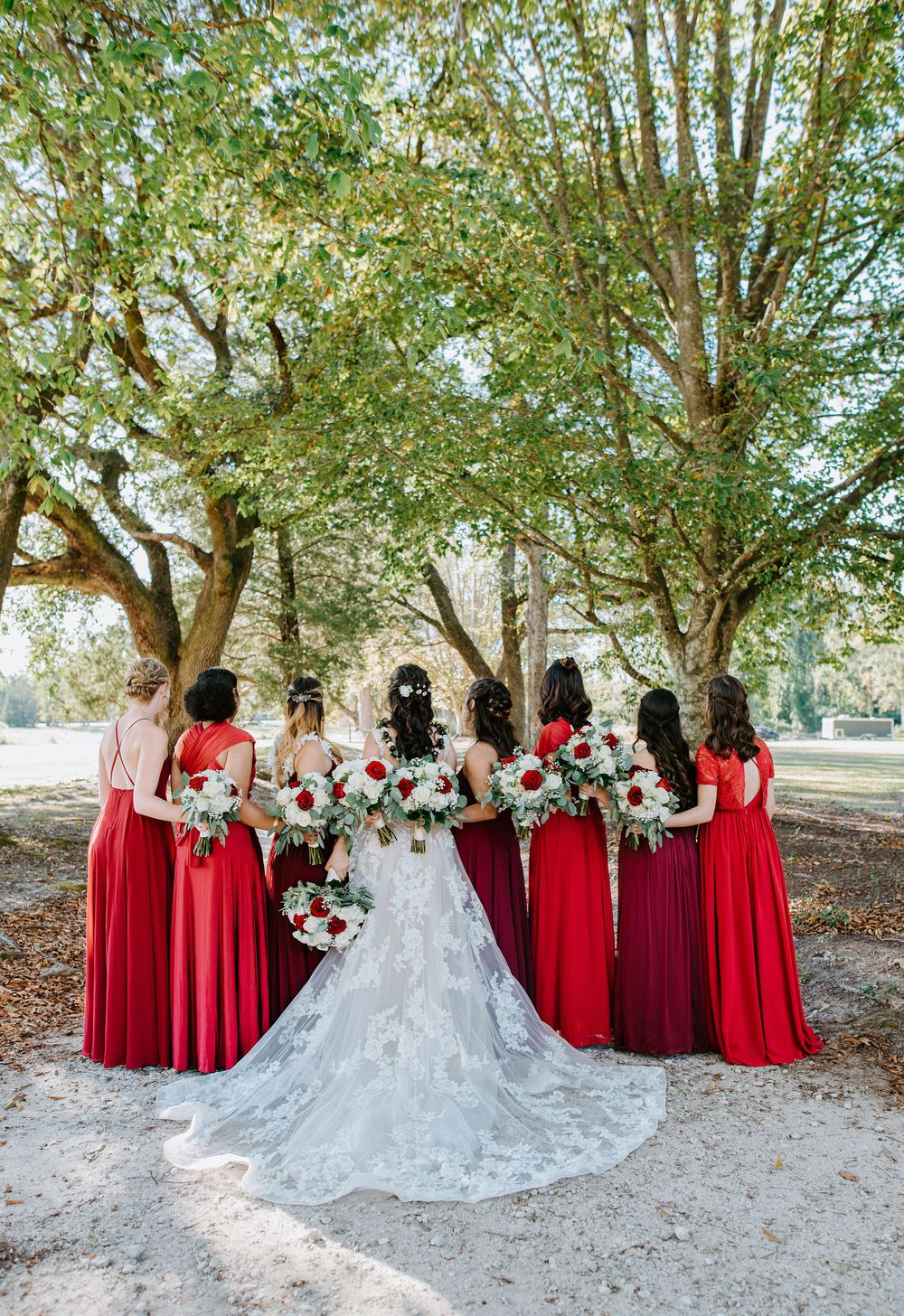 Bridal party in red dresses poses with bride in flowing white gown under tree canopy during outdoor photo session.