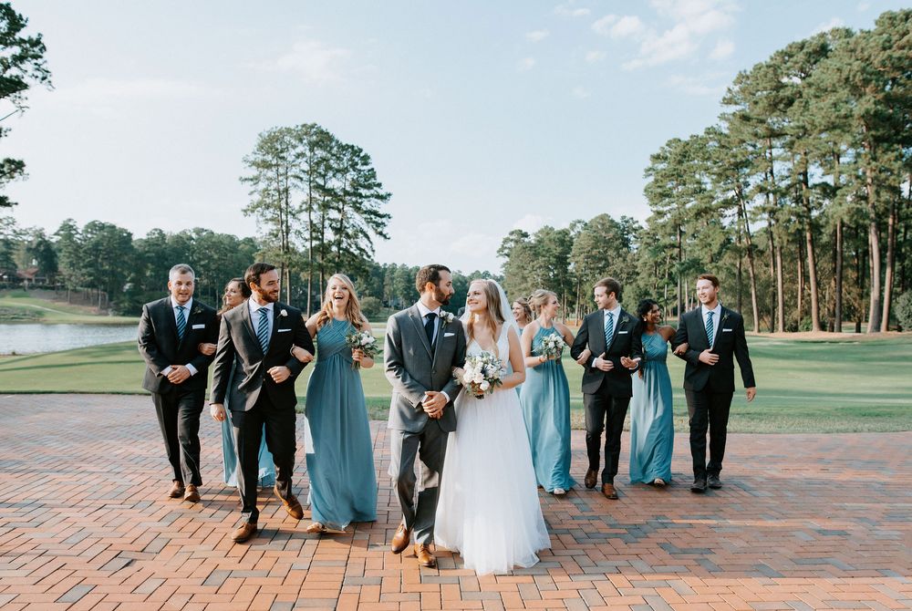 Wedding party in blue dresses and dark suits walking together on brick pathway at golf course with pine trees.