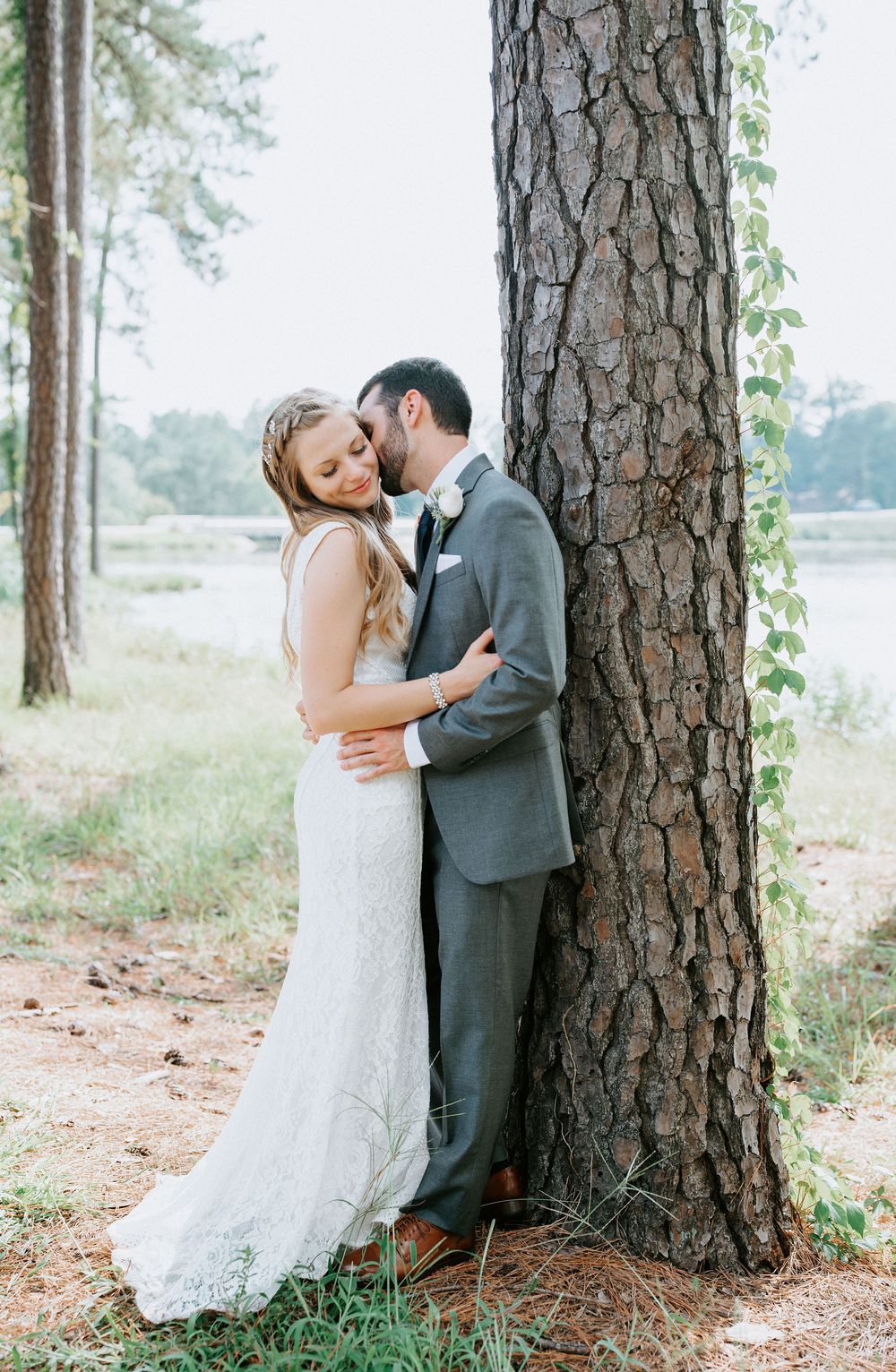 A romantic wedding couple embraces near a large pine tree trunk in an outdoor forest setting.