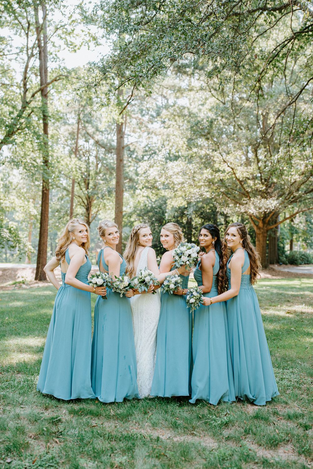 Bridesmaids in matching light blue dresses pose together in a line outdoors among trees.