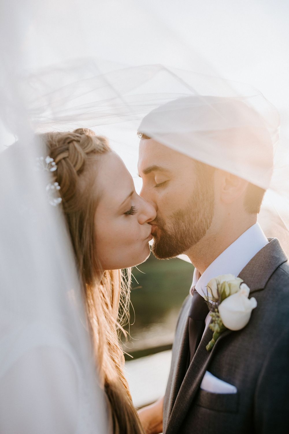 A romantic wedding couple shares an intimate kiss at sunset while wearing formal attire and white floral boutonniere.
