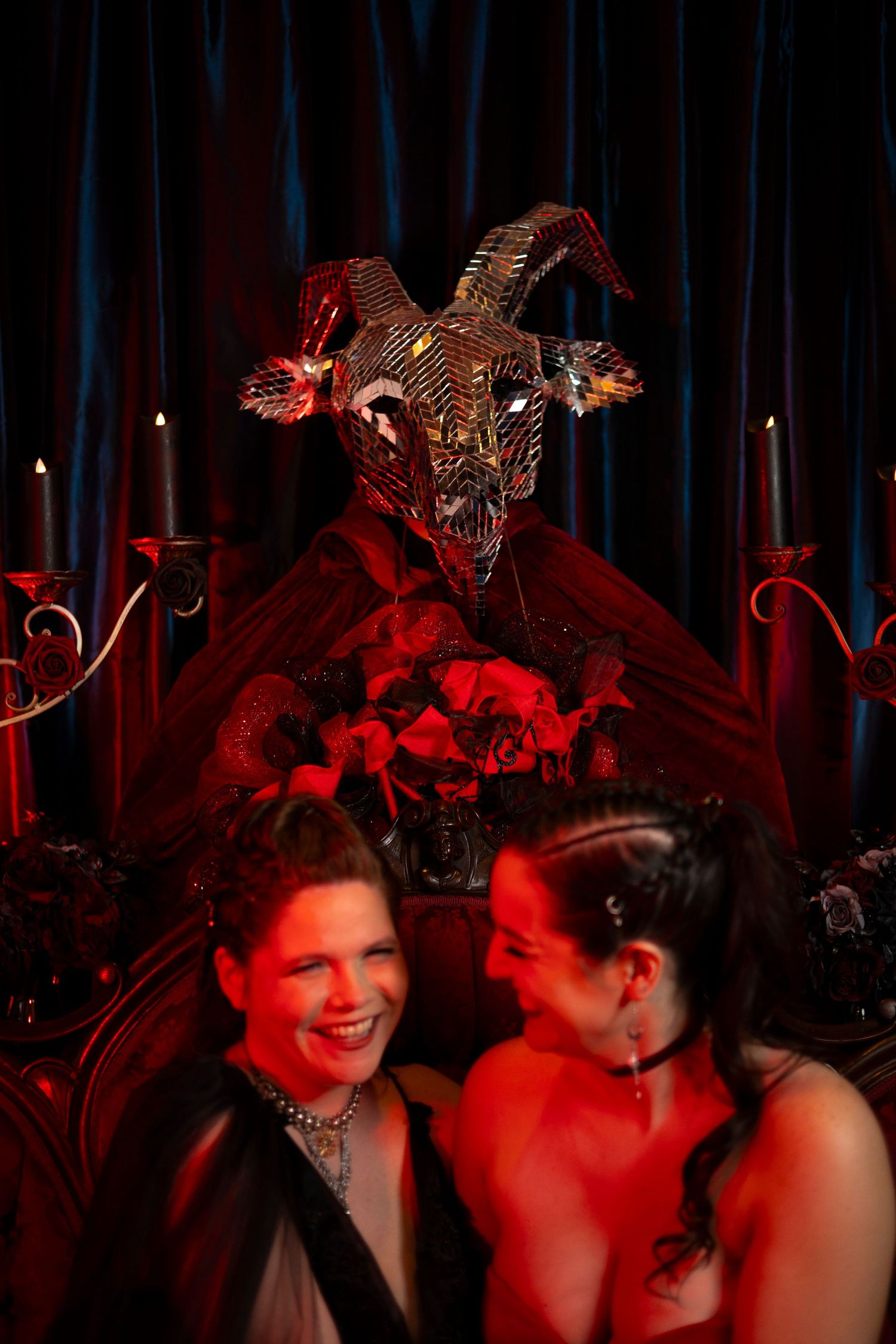 Two people in elegant red attire pose dramatically on a vintage sofa against dark damask wallpaper in moody lighting.
