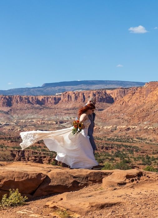 A bride in a flowing white dress poses dramatically on red rock formations in Utah's desert landscape at sunset.