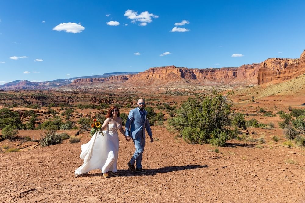 A couple walks hand in hand on a red dirt trail with towering sandstone cliffs and blue skies in Capitol Reef National Park.