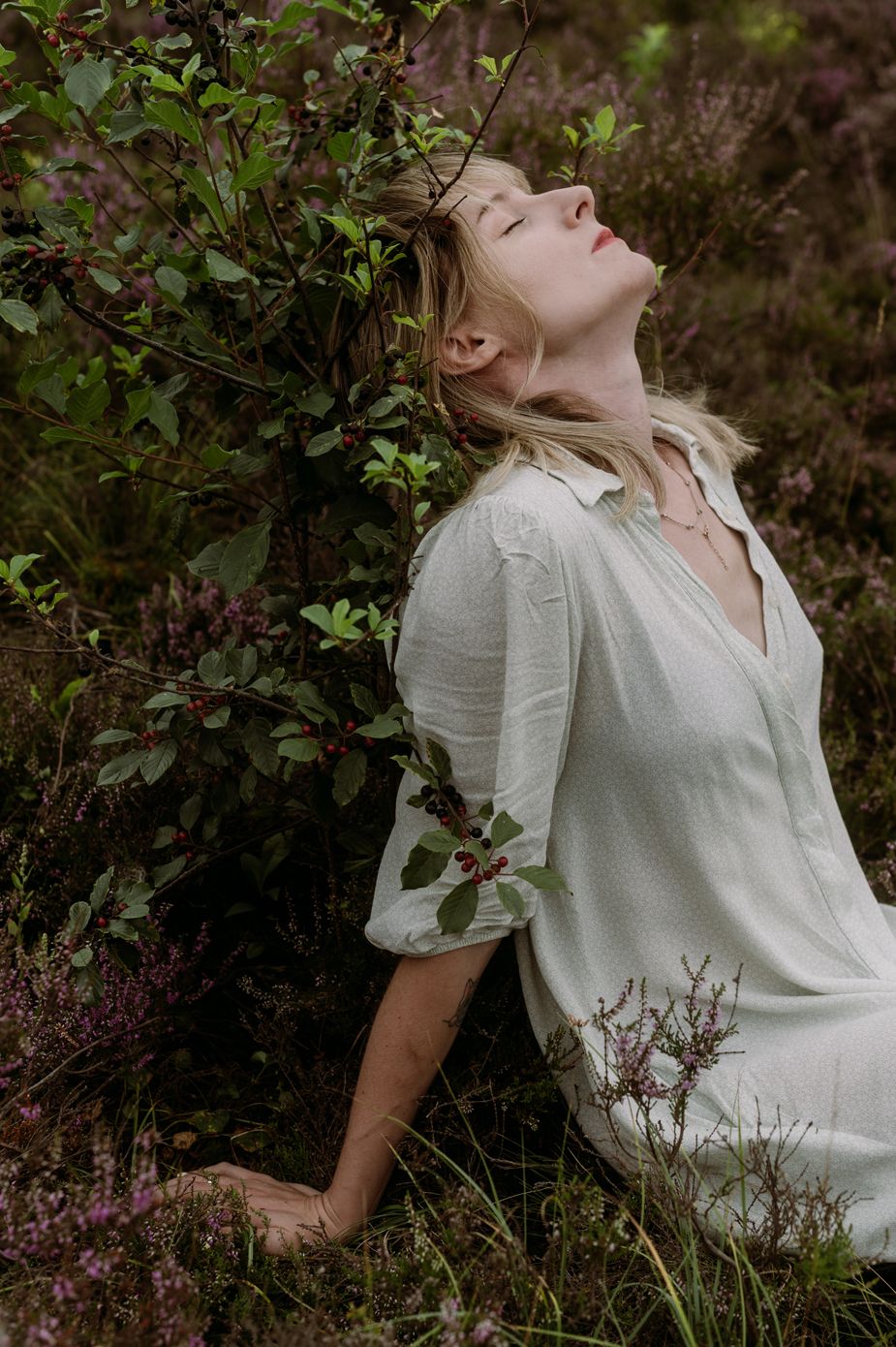 A person in a white vintage dress poses dramatically against a backdrop of purple wildflowers and green foliage.