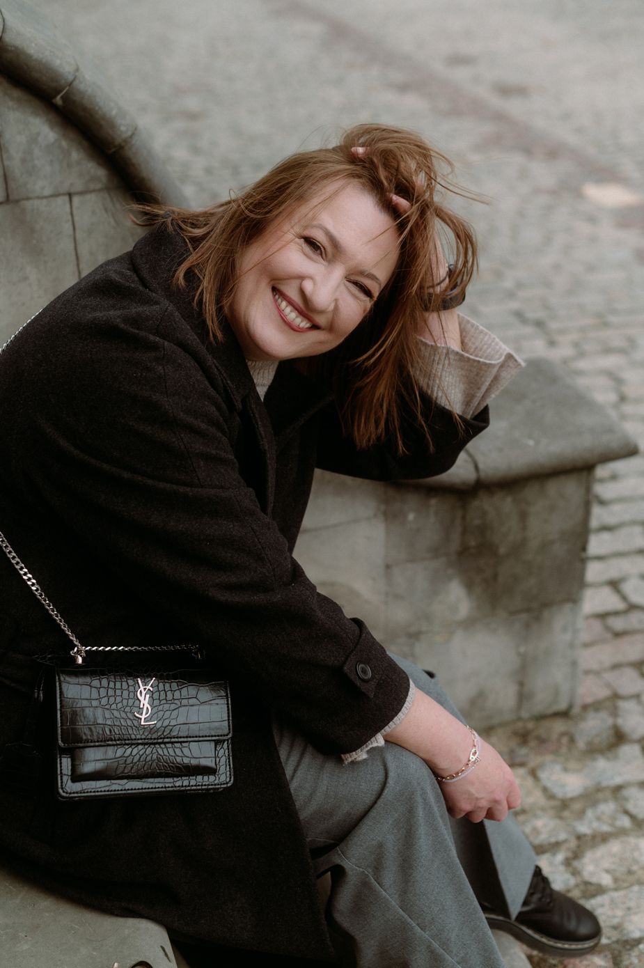 Woman in dark coat sits casually on stone steps with leather handbag in candid outdoor photo.