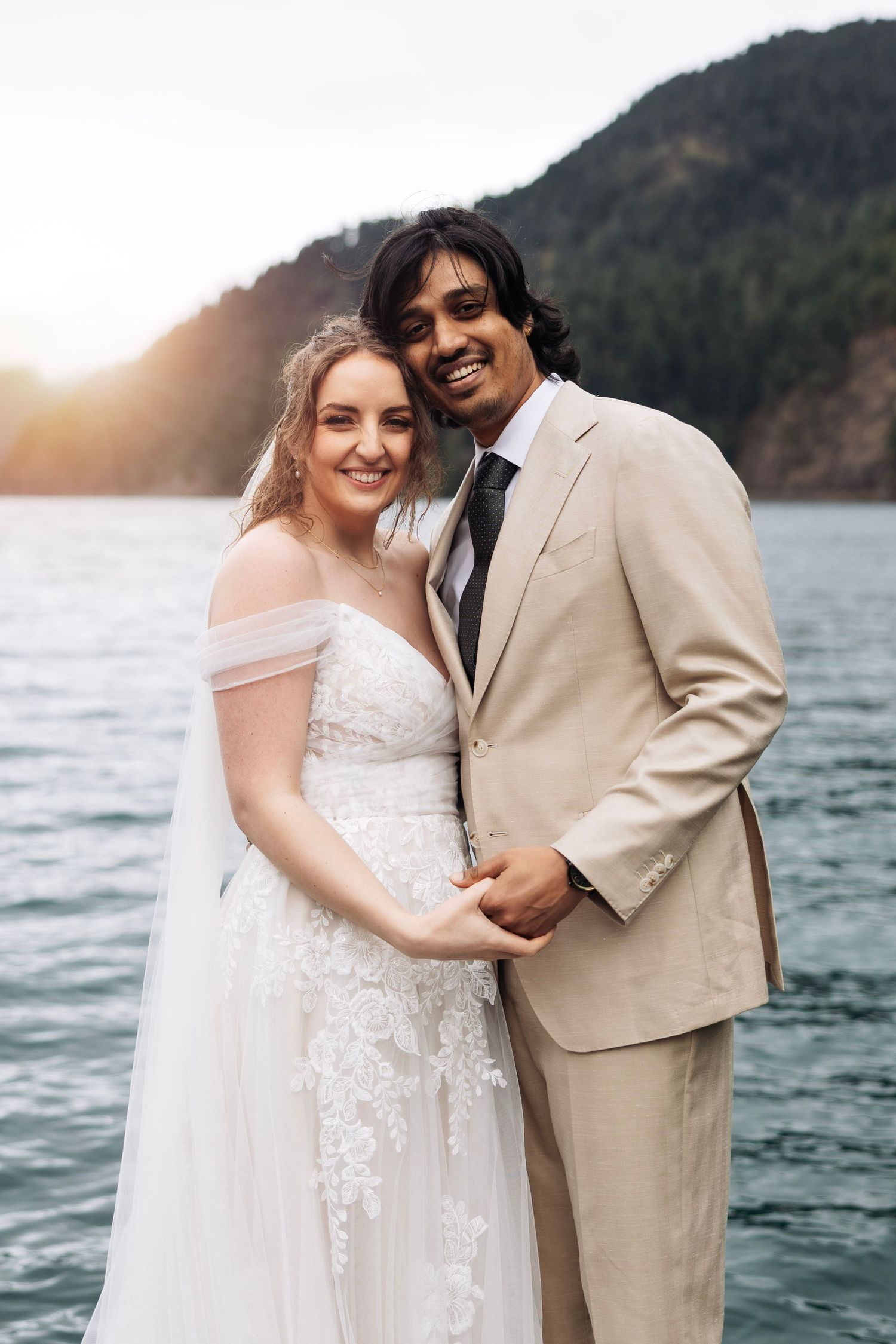 A couple in wedding attire embrace by a scenic lake with mountains and sunset in the background.