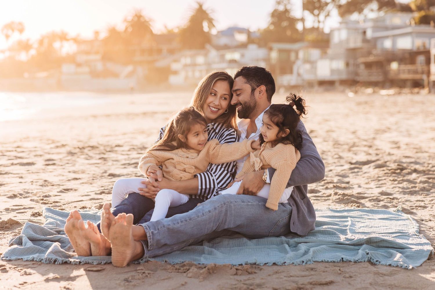 A family sits together on a blanket at the beach during golden hour sunset with coastal homes in the background.