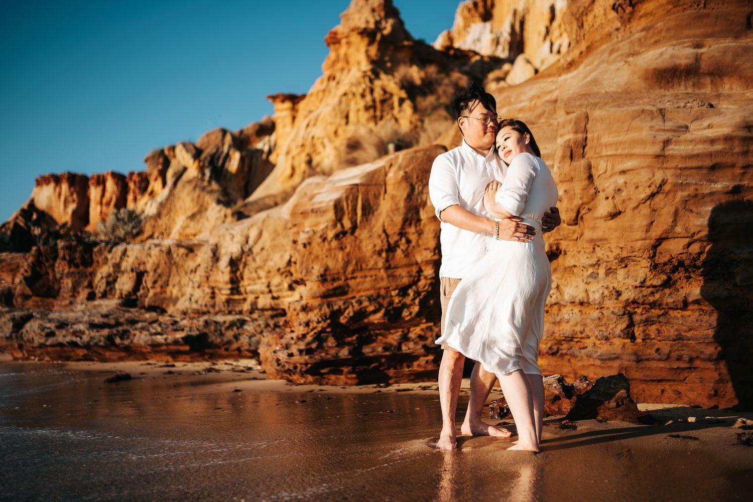 A couple in white outfits poses together against dramatic coastal rock formations at sunset taken by a Melbourne pre-wedding photographer.