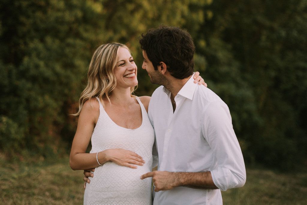A couple in white clothing shares an intimate moment outdoors during a maternity photo session in nature.