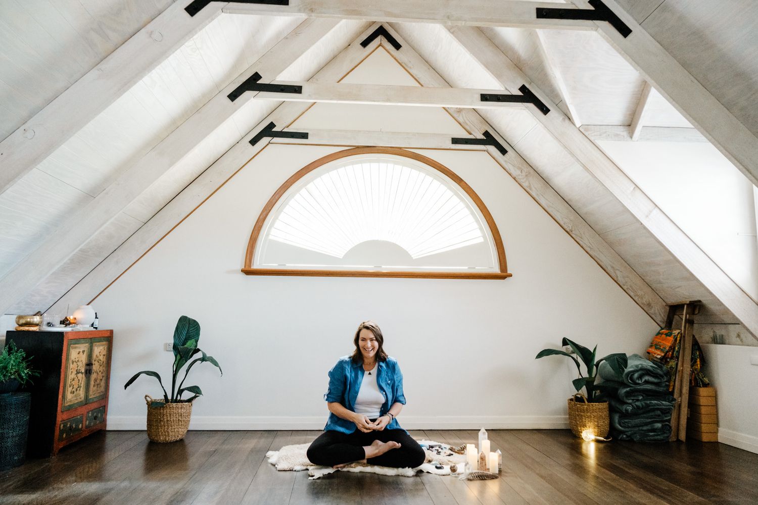 Person in casual clothing sits cross-legged on wooden floor with houseplants and decorative lighting in bright room.