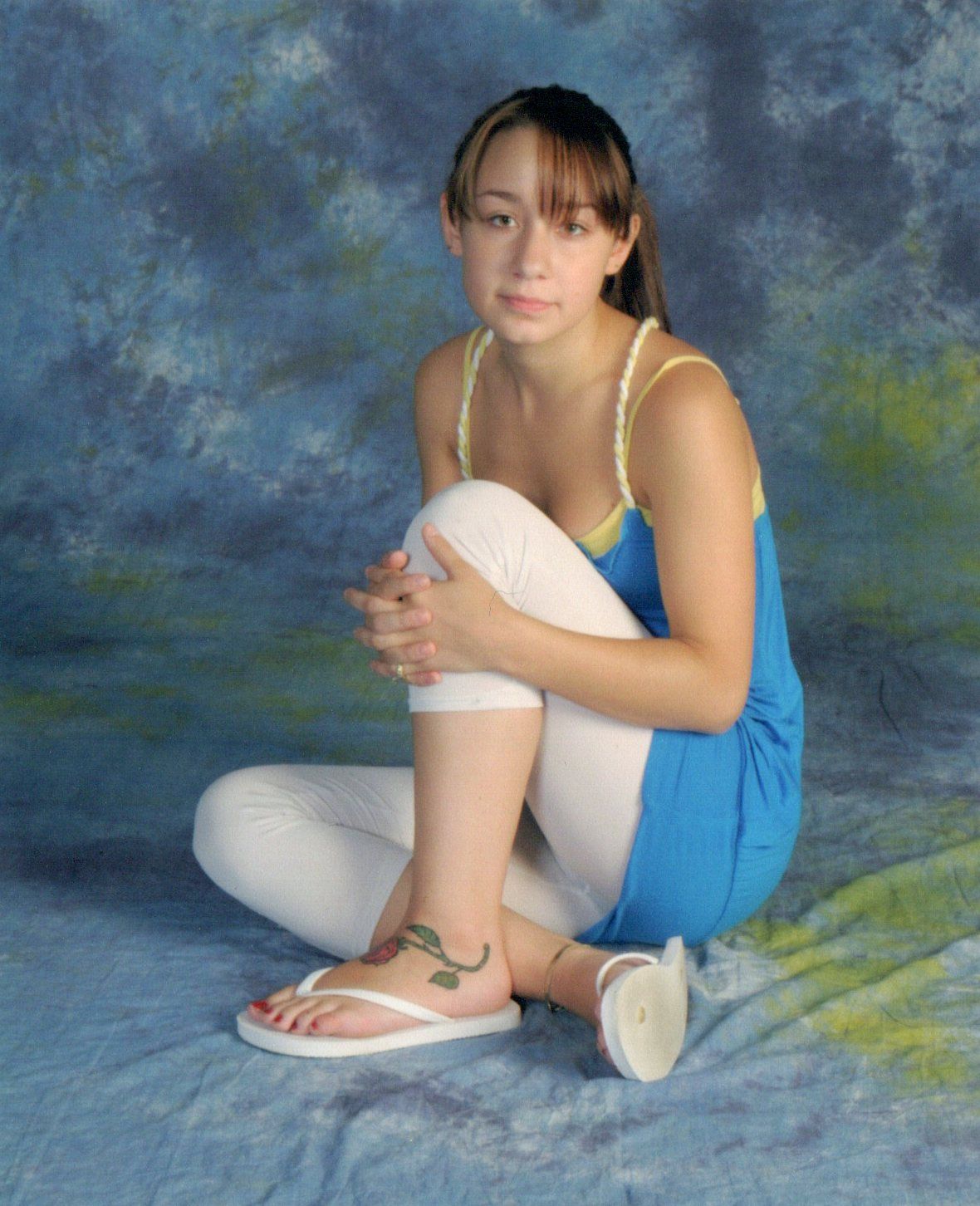 Person in blue tank top and white shorts sitting in relaxed pose against textured backdrop.