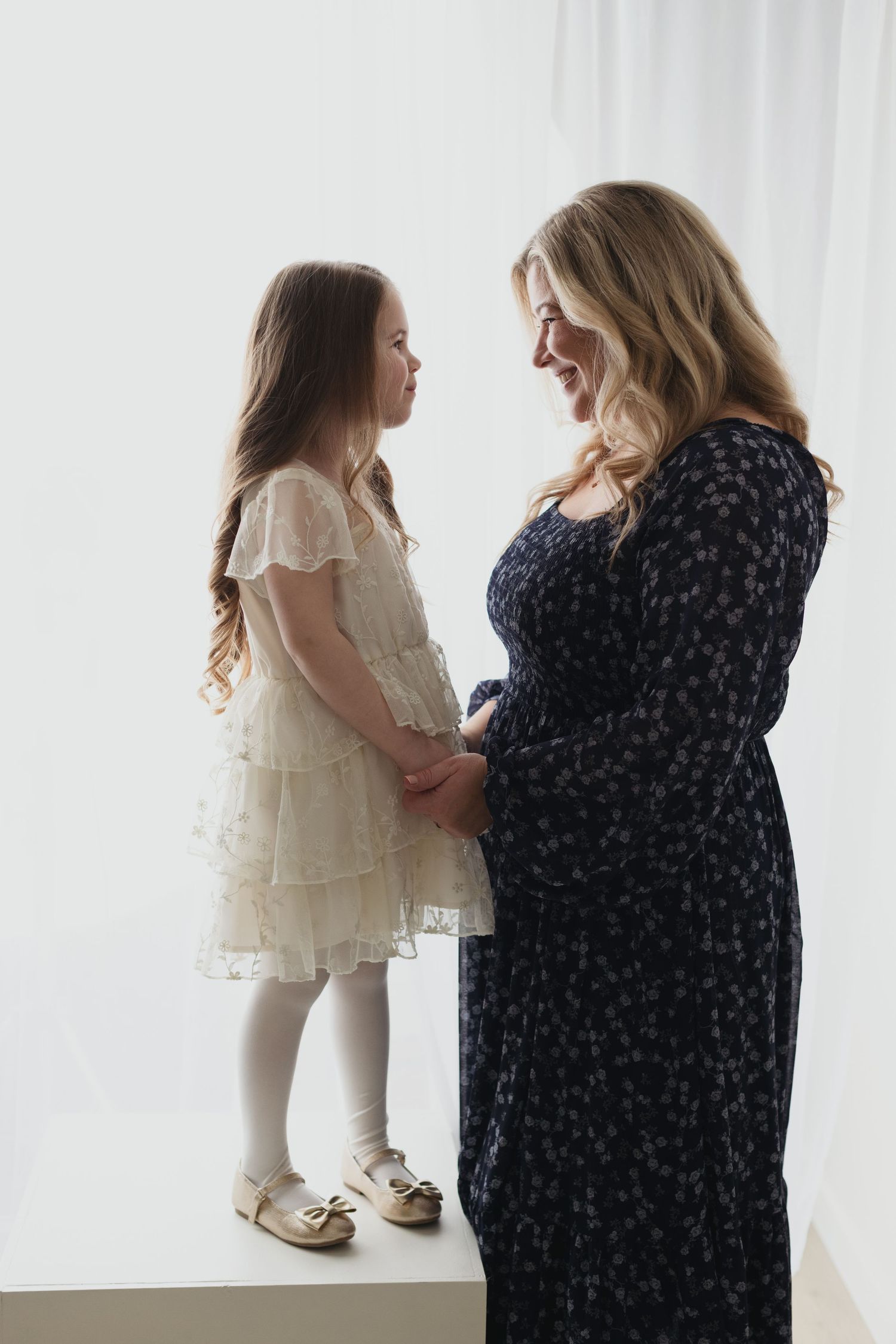 A touching moment between two people in contrasting dresses - one in white lace and one in navy floral - against a light backdrop.