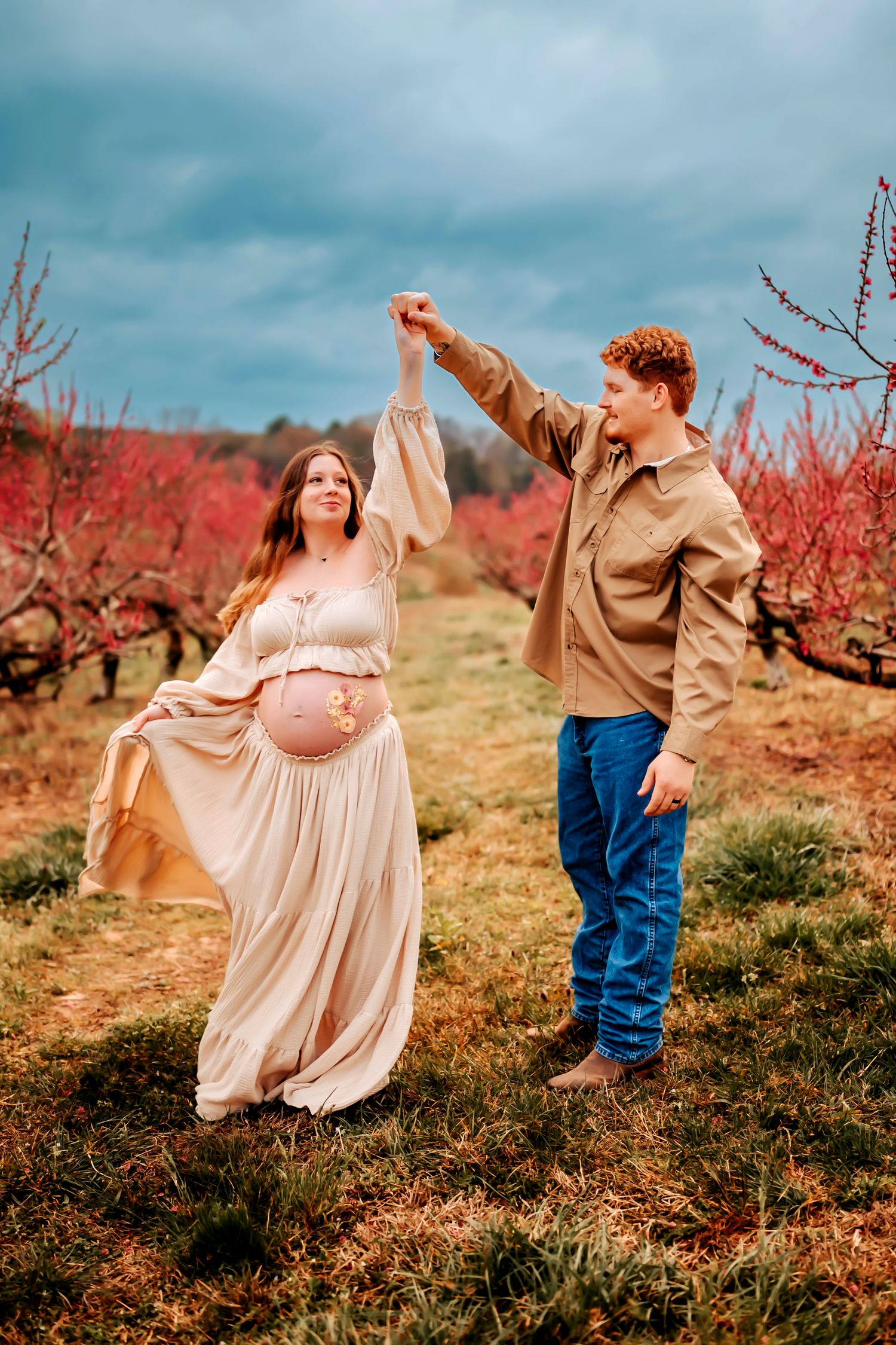 A couple dances playfully in a peach orchard during springtime with pink blossoming trees in the background.