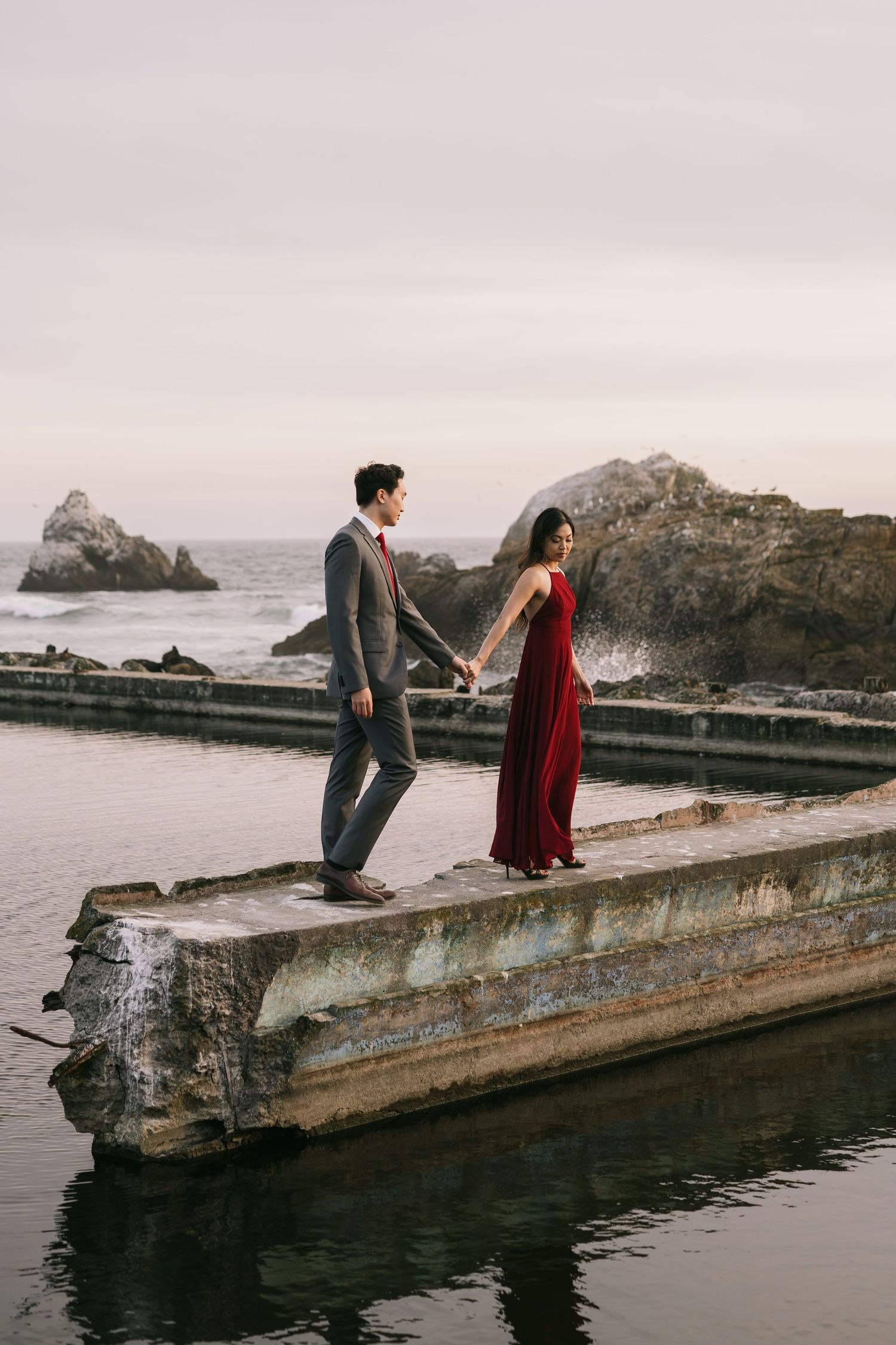 Couple walking hand in hand on a weathered concrete pier, woman in red dress, ocean rocks in background.