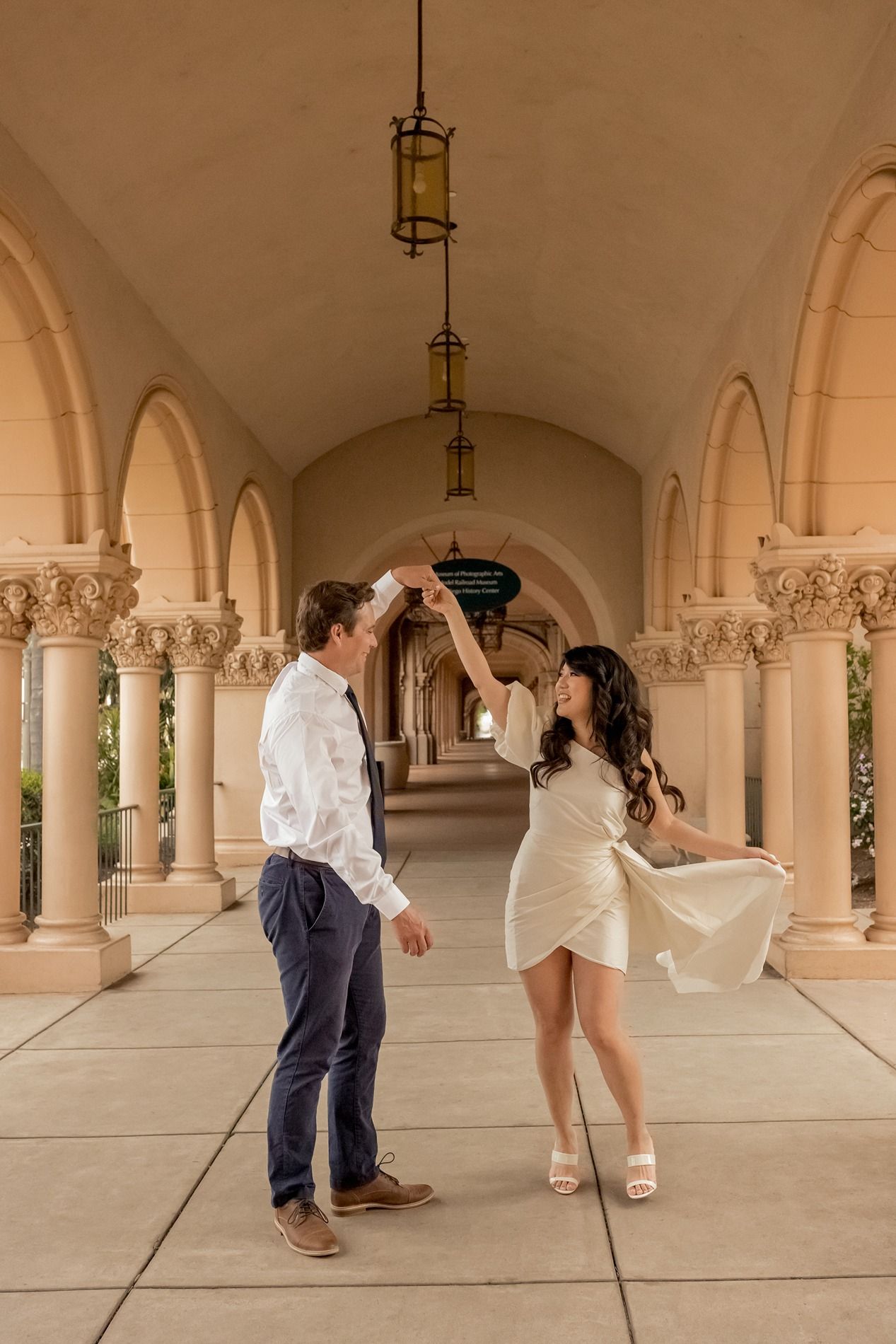 A couple dances playfully under an elegant stone archway with decorative columns and vaulted ceilings at Balboa Park in San Diego.
