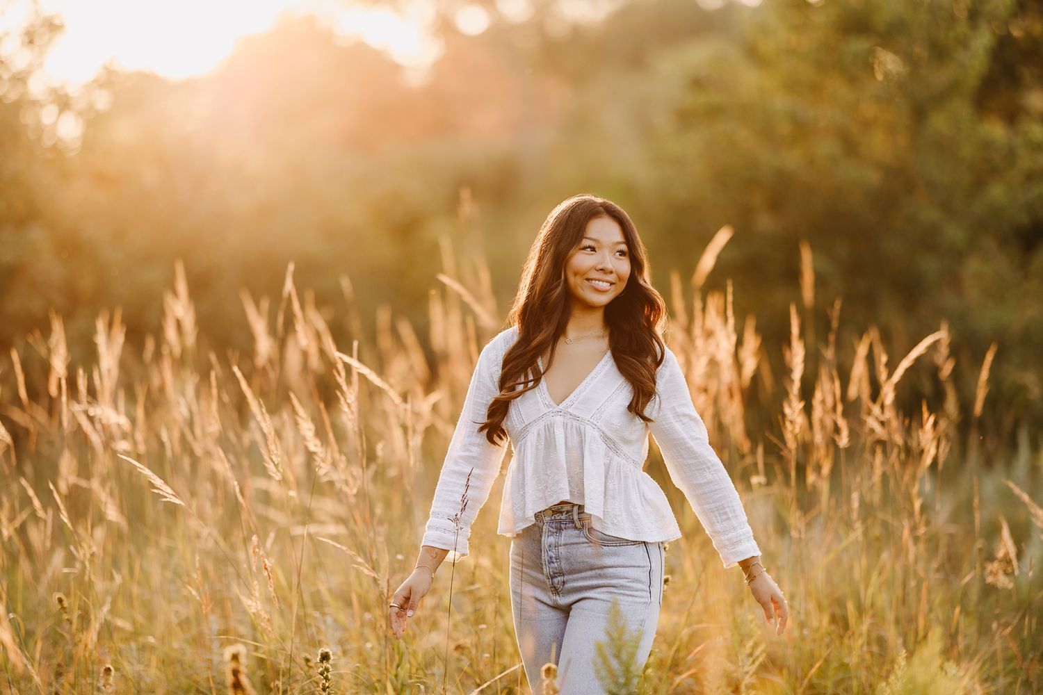 A person in a white flowing top and light jeans sits in a sunlit meadow with tall golden grass.