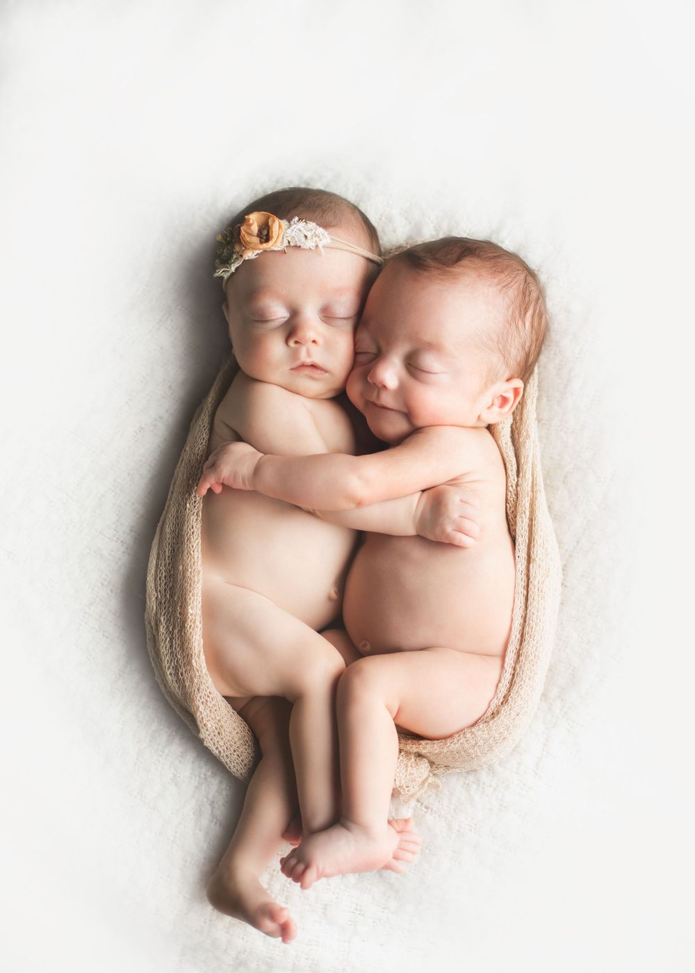 Two sleeping newborns cuddling together on soft beige fabric in a minimalist portrait setting.