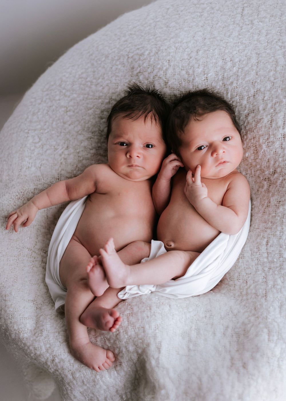 Twin babies lying together on soft white textured blanket.