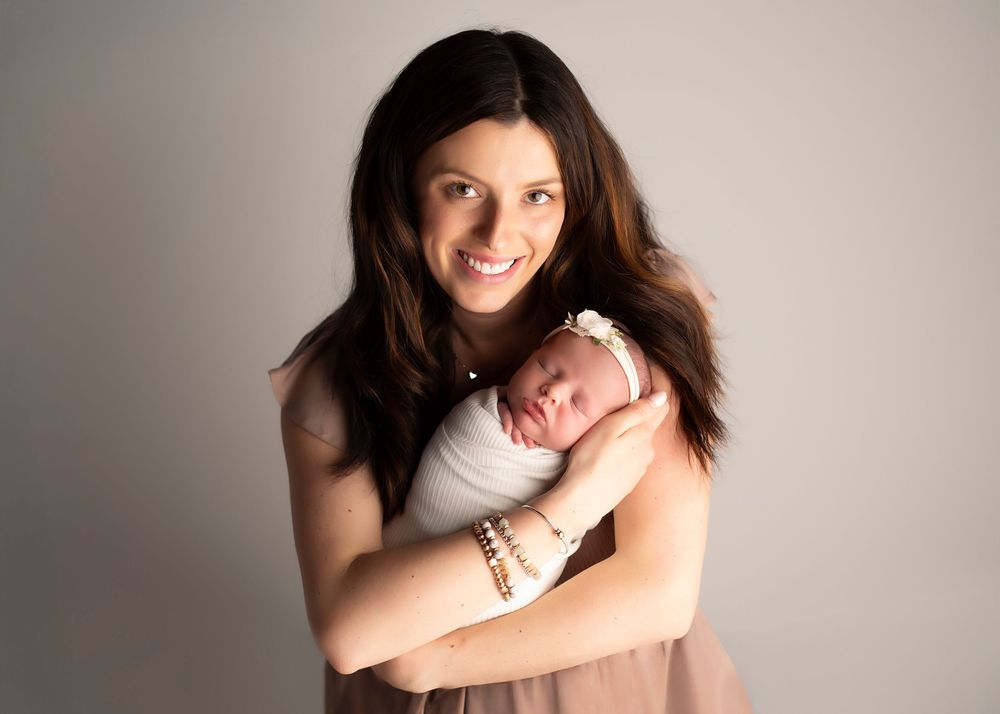 A tender moment captured as a woman in a pink dress cradles a sleeping newborn baby against a neutral backdrop.